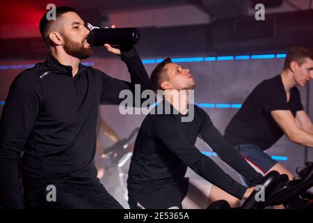 fit man takes a sip of water during working out in gym, cycling machine bicycle with friends Stock Photo