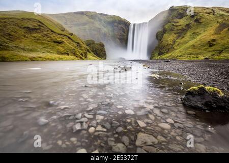 Iconic icelandic landscape with a stream flowing over pebbles in foreground and the Skogafoss waterfall, surrounded by green hills, in the background Stock Photo