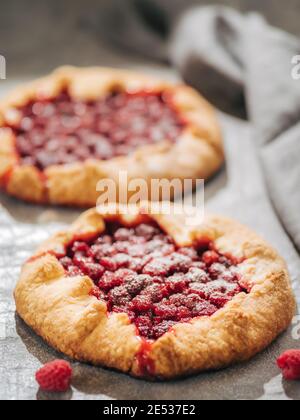 Two perfect raspberry galettes. Delicious rustic homemade tart with frozen or fresh raspberries on gray background. Beautiful round shape galettes with raspberries, copy space. Vertical. Shallow DOF Stock Photo