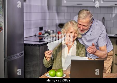 elderly man and woman managing family monthly budget together, positive married couple using computer banking application, counting bills in the kitch Stock Photo