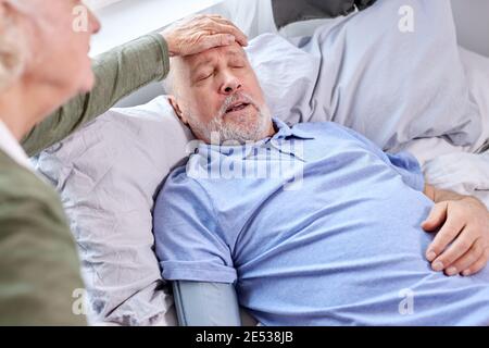 female checking fever temperature of senior man lying on bed. mature husband feeling symptoms of the flu while wife checking fever by touching forehea Stock Photo