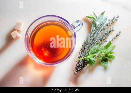 Picture of glass cup with tea, herbs and sugar on marble surface. Stock Photo