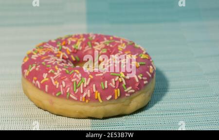 Pink sweet doughnut or donut with colored sprinkles on a blue background. Bitten off American bagel. Stock Photo