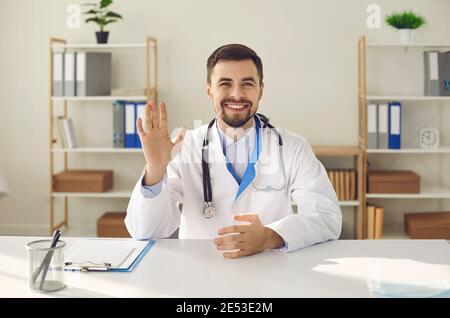 Happy doctor sitting at desk and waving hand, greeting patient before online consultation Stock Photo