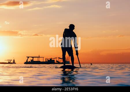 Active paddle boarder. Black sunset silhouette of young sportsman paddling on stand up paddleboard. Healthy lifestyle. Water sport, SUP surfing tour
