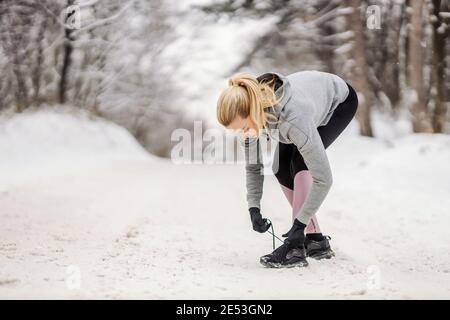 Sportswoman tying shoelace while standing on snowy path at winter day. Sportswear, healthy lifestyle, winter fitness Stock Photo