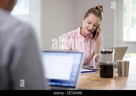 Business Couple Working From Home Sitting At Table During Pandemic Lockdown Using Mobile Phone Stock Photo