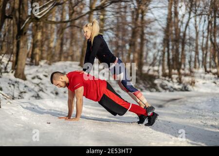 Sportsman doing push ups at winter while sportswoman standing in his backs. Relationship, winter fitness, healthy life Stock Photo