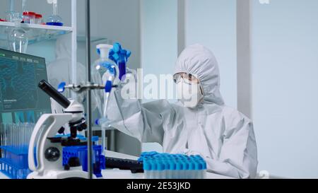 Medical scientist with coverall in laboratory room performs tests with blue liquid on test tube typing. Doctors examining vaccine evolution using high tech researching diagnosis against covid19 virus Stock Photo