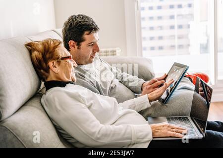 Side view of happy aged mother learning using laptop with help of adult son while sitting on sofa at home Stock Photo