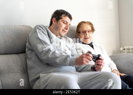 Adult male sitting on couch with elderly mother and teaching her using modern joystick for playing video game Stock Photo