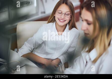 Funny cheerful teenager girl in white shirt and glass looking ahead near blonde woman Stock Photo