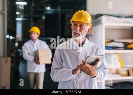 Picture of mature male manager with helmet on is head standing in warehouse with tablet in his hands. Looking away. Stock Photo