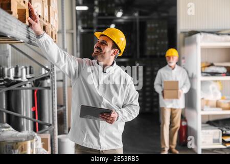 Picture of smiling young man warehouse worker counting boxes in warehouse. Holding digital tablet in his hands. Stock Photo