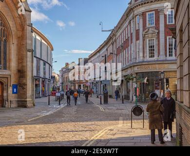 A winter afternoon in the city. Two people stop to talk on a corner as the sun throw light onto the cobbled street. Pedestrians and shops are in the d Stock Photo