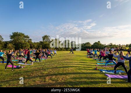A group participates in an outdoor yoga class at sunset in parkland, lit by the setting sun which casts shadows in patterns across the field. Stock Photo