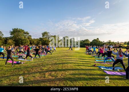 A group participates in an outdoor yoga class at sunset in parkland, lit by the setting sun which casts shadows in patterns across the field. Stock Photo