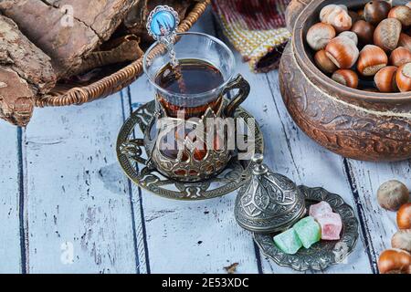 Black tea in a traditional glass cup with candies and a bowl of hazelnuts on blue wooden table Stock Photo