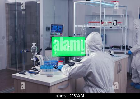 Scientist sitting at his workplace using computer with green screen. Team of microbiologists doing vaccine research writing on device with chroma key, isolated, mockup display. Stock Photo