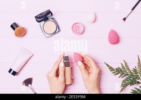 Makeup flat lay composition female hands holding bottle of foundation and sponge applicator among makeup brushes and cosmetic products on pink backgro Stock Photo