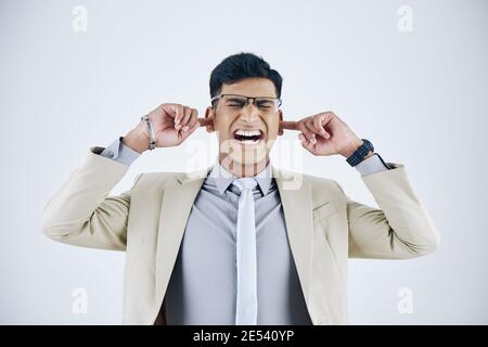 Annoyed irritated and stressed out young Indian businessman plugging ears with fingers and screaming Stock Photo