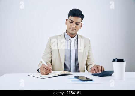 Smiling handsome young businessman sitting at office desk, making calculations and writing in planner Stock Photo