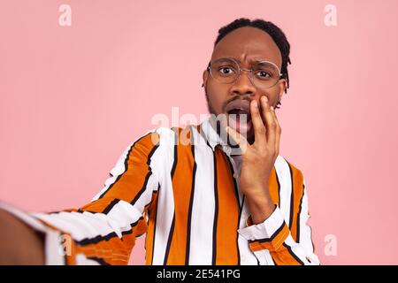 Surprised shocked afro-american man with dreadlocks in eyeglasses stylish striped shirt looking at selfie camera with astonishment, widely opening mou Stock Photo
