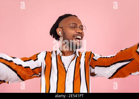 Cheerful happy bearded afro-american man with dreadlocks in eyeglasses and earphones looking at camera and smiling, making selfie, posing. Indoor stud Stock Photo