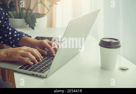 Young woman using laptop, communicates on internet with customer in home garden/greenhouse, reusable coffee/tea mug on table. Cozy office workplace, r Stock Photo