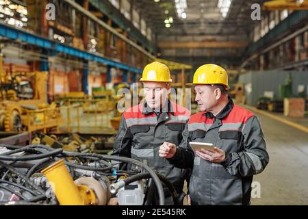 Two workers in helmets using tablet pc and discussing the work of machine equipment in the plant Stock Photo