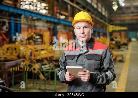 Portrait of mature manual worker in helmet and workwear looking at camera while using tablet pc in the factory Stock Photo