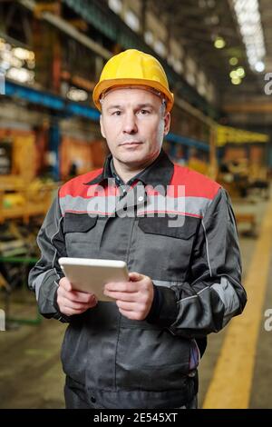 Portrait of mature manual worker in helmet looking at camera while working online on tablet pc in the factory Stock Photo