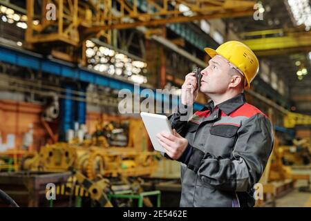 Mature foreman in helmet communicating on a wireless radio with his colleagues and using tablet pc in the plant Stock Photo