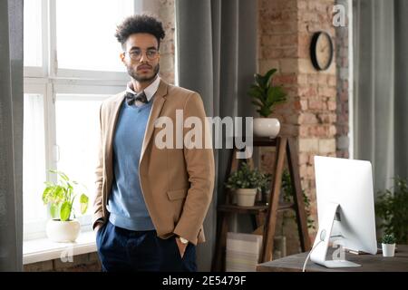Young designer in stylish suit standing near the window and thinking about something while working at office Stock Photo