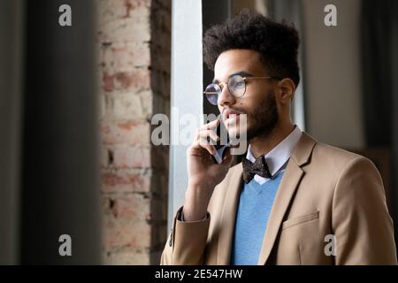 Young man in eyeglasses looking through the window during his conversation on mobile phone Stock Photo