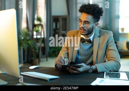 Young businessman sitting at the table and looking at computer monitor he making notes during business meeting online Stock Photo