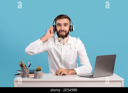 Bearded male helpline operator with headphones and laptop answering customer's call at desk over blue studio background Stock Photo