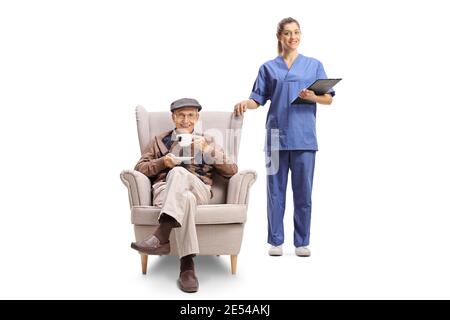 Female nurse holding a clipboard and standing next to an elderly man in an armchair with a cup of tea isolated on white background Stock Photo