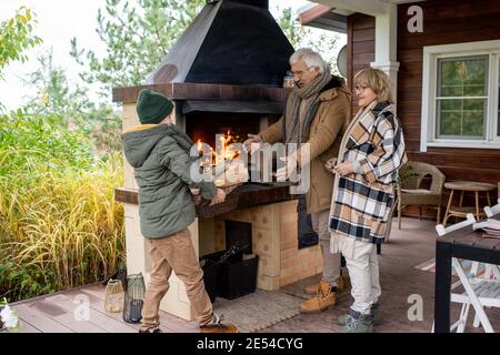 Happy senior couple in warm casualwear standing by fireplace of their country house and looking at grandson carrying box with firewood Stock Photo