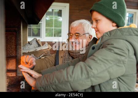Happy senior man and his grandson standing by fireplace with burning firewoods while getting warm on cool autumn day by country house Stock Photo
