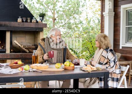 Happy mature woman in eyeglasses and warm casualwear looking at her husband while both having tea with homemade snacks against fireplace Stock Photo