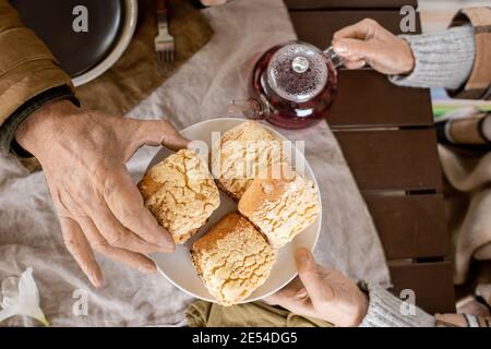 Hand of aged man taking fresh homemade bun from plate held by his careful wife over served table while both having breakfast Stock Photo