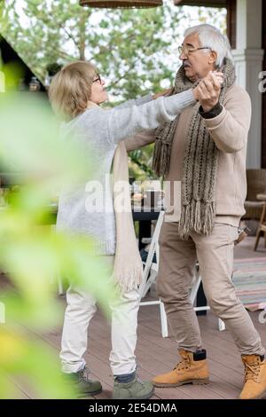Affectionate senior couple in warm stylish casualwear dancing on wooden floor of patio by their country house against served table Stock Photo