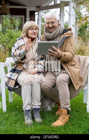 Elderly husband and wife with digital tablet communicating in video chat while sitting on bench on green lawn in front of their country house Stock Photo