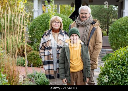 Happy youngster in casualwear and his affectionate grandparents looking at you with smiles while standing in front of camera in the garden Stock Photo