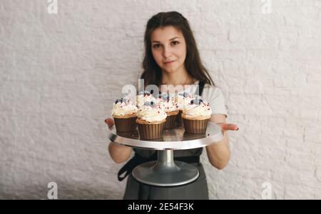The cook girl holds a tray with beautiful cupcakes decorated with cream and berries. Stock Photo