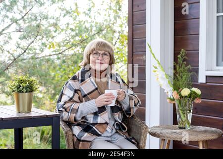 Retired blond female in eyeglasses and warm stylish checkered coat holding mug with hot tea while sitting in armchair by country house Stock Photo