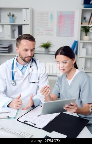 Two young colleagues in medical uniform sitting by desk in medical office and discussing online information on tablet display during presentation Stock Photo