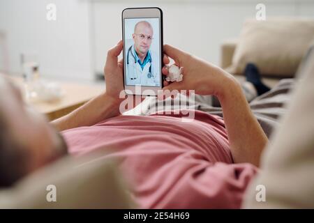 Confident mature male doctor on smartphone screen giving medical recommendations to young sick man with handkerchief lying on couch Stock Photo