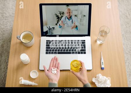 Mature male doctor on laptop display giving medical recommendations to sick female holding pills that she takes and showing them to him Stock Photo